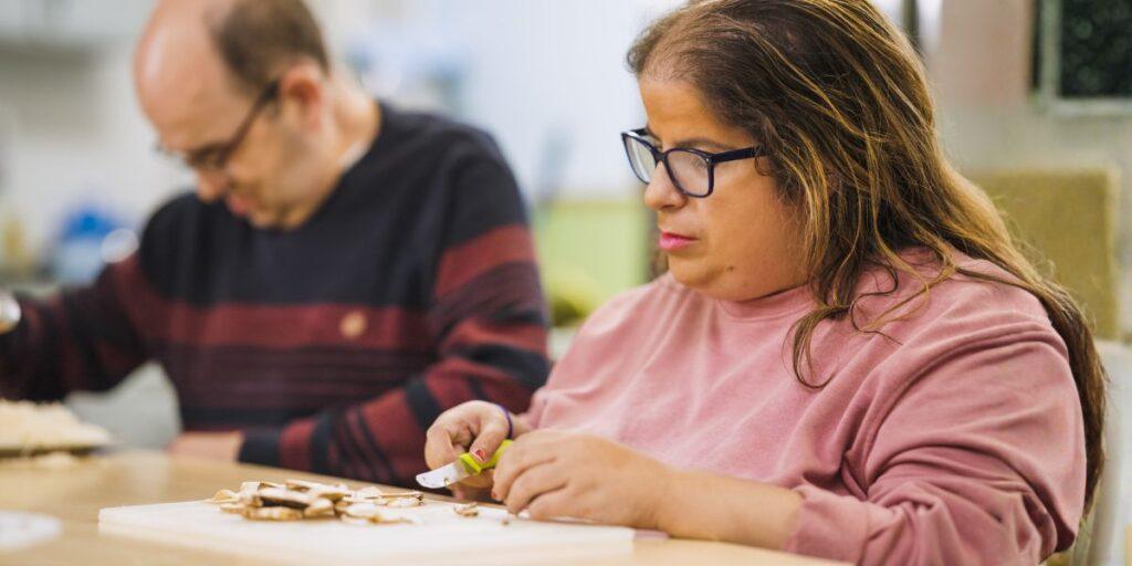 A woman using a knife to slice mushrooms at a table, with a man preparing food beside her.